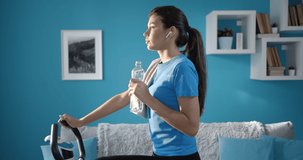Side view of confident young woman in sportswear drinking water on stationary bike in living room with grey and blue interior colors. Concept of health care, habits and motivation.