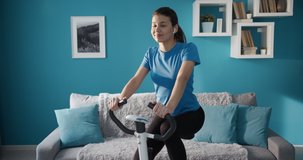 Happy active girl with dark hair in sportswear listening to music in wireless headphones while training on exercise bike. Sporty brunette doing daily workout at home.