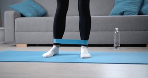 Close up of young fitness lady doing legs exercises with elastic bands while standing on yoga mat in white socks. Healthy woman strengthening her muscles with sport equipment at home.