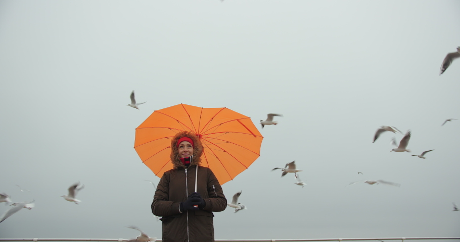 Woman in parka with orange umbrella in heart shape enjoying sea view with seagulls in storm. Overcast weather.