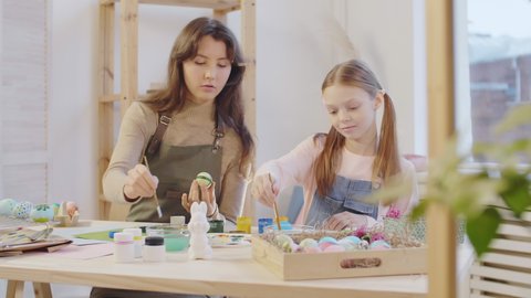 Medium shot of young woman and cute preteen girl decorating Easter egg together. They are using small brushes and drawing pattern with gouache paint