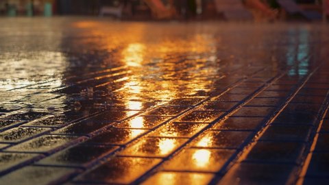 Rain drizzles along wet sidewalk, city street lights reflected in puddle at evening, drops fall and splash, pavement is filled with water. Blurred background, shallow depth of field.