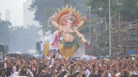 A giant Ganapathi idol driven in the street for immersion during the Ganapathi festival. A crowd of people are happily dancing along the street in the rain, Mumbai, India (August 2019)