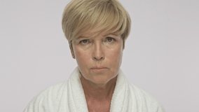 A sad upset adult woman with short blond hair wearing white housecoat is showing her tooth pain isolated over white background