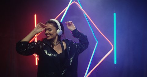 Caucasian cheerful young brunette woman in headphones and latex black coat dancing and listening to music at neon tubes light on background. Female meloman concept.