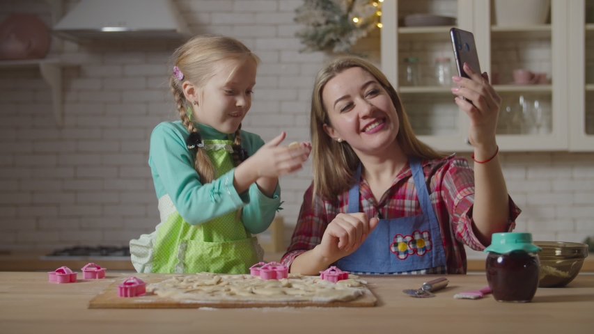 Cheerful mother and lovely preadolescent daughter in aprons live streaming with cellphone during cooking in domestic kitchen. Family making online video chat with smartphone while preparing cookies.