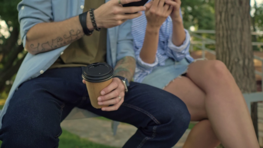 Close-up view of a young smiling couple is using smartphones while sitting on the bench with electric kick scooters at a park landscape