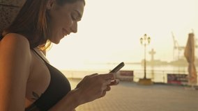 Side view of Cheerful pretty brunette sports woman using smartphone and enjoying this moment near the sea outdoors