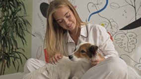 A nice young woman wearing white pajamas is petting her dog while laying on a bed at the bedroom