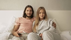 A positive young couple man and woman are watching television using the remote control in the bedroom at home