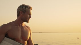 Cool bearded muscular surfer holding surfboard and walking on sunset near the sea