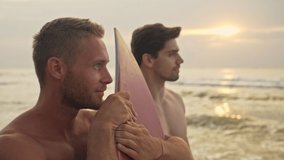 Side view of Two cool muscular handsome surfers standing with surfboards and looking away near the sea