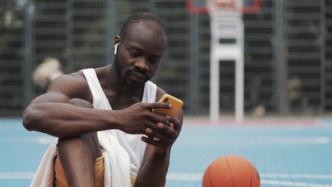 Young Musculy Afro American Guy in Earphones Sitting on the Ground , Using his Smartphone and Smiling at Street Basketball Court. Healthy Lifestyle. Tecnology and Sport Concept.