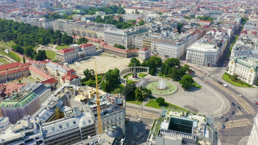 Vienna Austria Monument to Soviet soldiers. An aerial shot reveals a dense European cityscape under a bright, clear sky. The scene is dominated by the Schwarzenbergplatz, a large circular plaza featuring a central fountain and the prominent Soviet War Memorial with its tall, slender obelisk and surrounding colonnade. The plaza is bordered by manicured lawns and mature trees. To the left, a large, open square with sandy-colored pavement adjoins a complex of buildings with distinctive red-tiled roofs. A yellow construction crane is visible in the lower-left corner. The vast background extends into a continuous grid of multi-story buildings with light-colored facades and red roofs, interspersed with patches of green parks. The camera slowly pulls back and pans right.