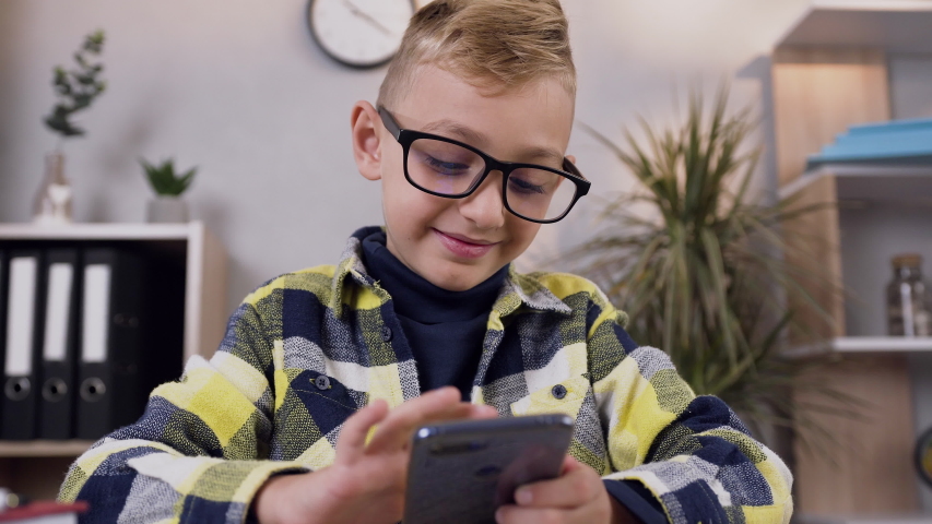 Slow motion of good-looking joyful modern blond 10-aged schoolboy in eyeglasses which browsing information on mobile screen
