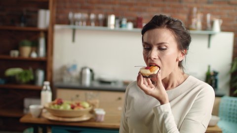 Portrait of enjoying girl biting dessert at home in slow motion. Closeup satisfied woman looking at camera with fancy cake in hand. Attractive brunette girl eating cake in home kitchen.