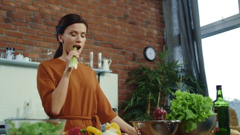 Beautiful girl eating fresh celery at home. Satisfied female tasting juicy celery. Young woman getting idea during cooking healthy food in kitchen. Fresh vegetables on kitchen table.