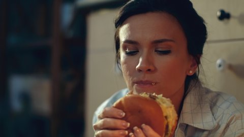 Closeup hungry girl eating big burger on kitchen floor. Portrait of young woman drinking red wine evening at home floor. Joyful woman enjoying fast food dinner and wine at home in slow motion.