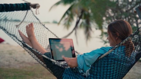 Young woman freelancer lying in a hammock with a laptop on the tropical beach at the sunset time.