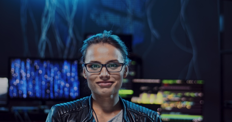 Close up of the young charming Caucasian girl hacker in glases looking straight to the camera and smiling cheerfully while sitting in the dark server room with computers on the digital world. Portrait