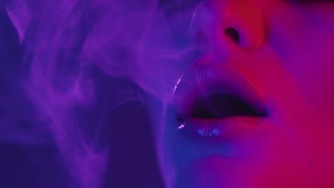 Colorful portrait of beauty sexy model girl in neon, blowing smoke of electronic cigarette or vape. Focused close-up of young woman face with dark make-up in UV pink and blue lights. Slow motion