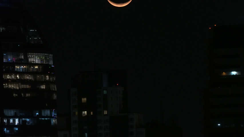 Crescent Moon time lapse setting over a modern city at night in between high rise buildings. 