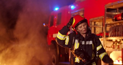 Portrait shot of the tired Caucasian young woman fire fighter taking off helmet and walking out from fire at the red truck. Outside late at night.