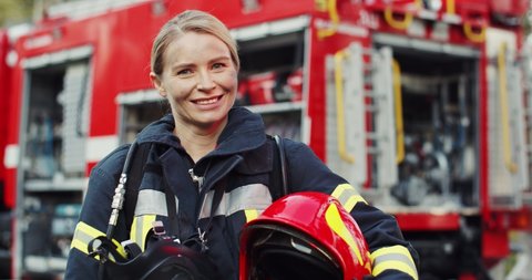 Portrait shot of the happy Caucasian young woman firefighter with the helmet in hand smiling to the camera after fire at the red truck. Outside.