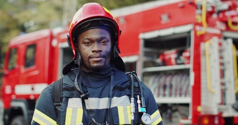 Portrait shot of the African American joyful firefighter taking off helmet and smiling to the camera at the big red truck after fire.
