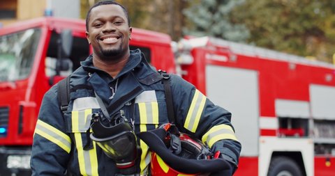 Portrait of the young joyful good looking African American fireguard taking off helmet and looking to the camera with a smile at the fire truck. Outdoors.