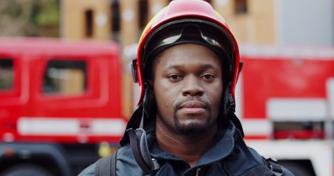 Portrait of the happy and handsome African American young fireman in helmet smiling cheerfully to the camera on the fire truck background.