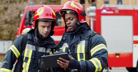 Caucasian and African American firemen in full equipped clothes standing outside at the fire truck with a plan in hands and deciding what to do for extinguishing fire.