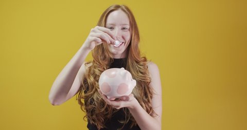 A redhead young woman with a pink pig coin bank, money box, for banking, saving, deposit. Yellow background.