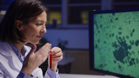 Side view zoom in of middle aged female biochemist in lab coat smelling red liquid in test tube sitting at desk in laboratory, magnified microorganisms on her computer screen