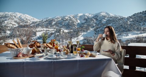 Young girl having a luxurious breakfast with various fruits and snacks, drinking her morning coffee and enjoying view of mountains - luxury lifestyle concept 4k footage