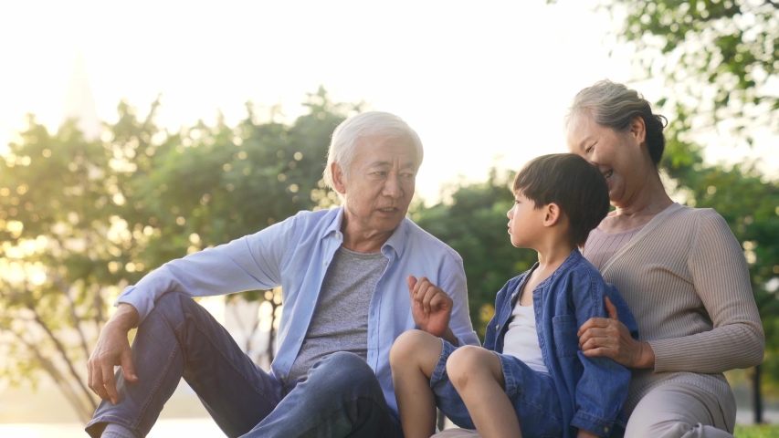 Happy senior asian couple sitting on grass having fun with grandson outdoors in park