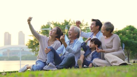 happy three generation asian family sitting on grass outdoors taking a selfie