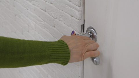 A female hand opens the white door in the white room by the doorknob and closes the door behind itself