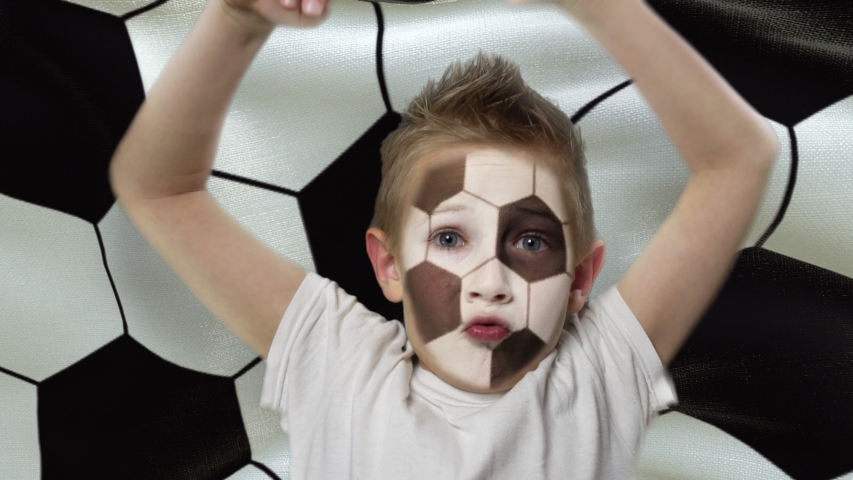 Football fan. Joyful fan on the background of the flag with a ball print. Happy boy with a painted face. The young fan rejoices in the victory of his beloved team. Success. Victory. Triumph.