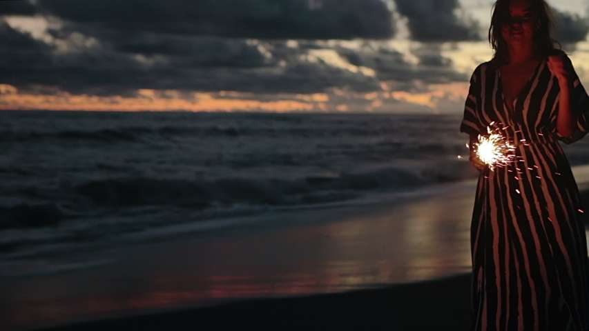 Close up portrait of young beautiful woman at sunset on the beach. Pretty girl holding sparkling fireworks in slow motion. Holiday with fireworks. City people travel. Business woman at vacation.