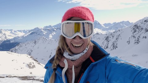 Happy young woman on ski slopes taking a selfie portrait on top of snowcapped mountains in winter enjoying ski season vacations. Girl having fun in Switzerland sharing selfie to friends video