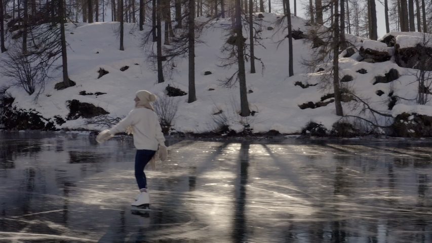 Tracking shot, woman ice skating on frozen lake in winter. Female on ice skates low later drone shot woman ice skating. Winter activity in the Alps 