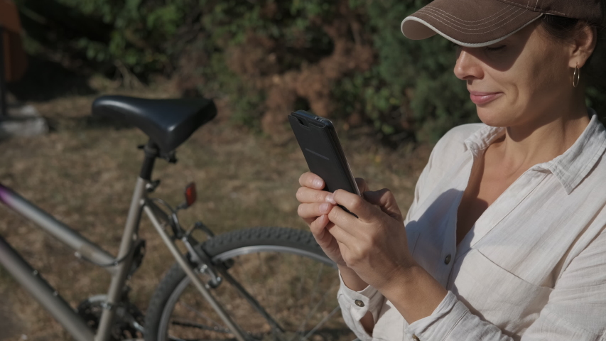 Have a rest after cycling. Happy woman sit and type a message on her telephone by the bicycle.