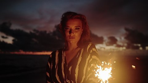 Close up portrait of young beautiful woman at sunset on beach. Pretty girl dancing in with sparkler at sunset in slow motion. Holiday with fireworks. City people travel. Business woman at vacation.