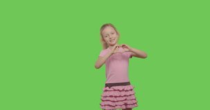 Close-up of smiling child girl making sign heart shape by hands posing at green screen studio background. Chroma Key 4k raw video footage slow motion 60 fps