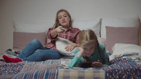 Charming mother watching tv, changing tv channels with remote control and cute preadolescent daughter watching video content online on mobile phone while family relaxing on bed in the evening.