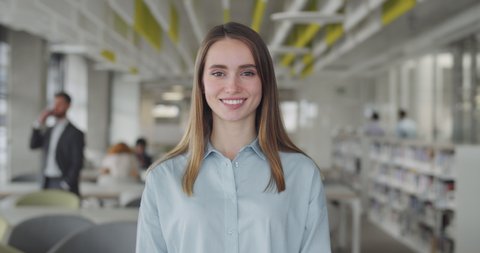 Smiling caucasian girl intern standing at modern office coworking space. Portrait of young female office manager looking to camera and having good mood. Blurred office background.