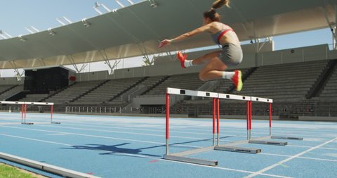 Rear view of a Caucasian female athlete practicing at a sports stadium, hurdling on running track, slow motion. Track and Field Sports Training in Stadium.