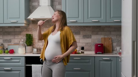 Happy Pregnant 20s Woman Mum Dancing in Home Kitchen. Funny Expectant Mother Listening Music and Sing Song at Home. Cheerful Happy Charming Woman Fun and Expressive Preparing Lunch. Concept of Family