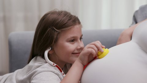 Curious Little Girl Plays at Doctor with Pregnant Female. Adorable Caucasian Daughter Examines a Pregnant Mom Belly with Toy Medical Equipment. Charming Kid and Mother have Fun Hospital Game. Closeup